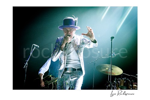 Horizontal color image of Gord Downie of The Tragically Hip singing into a microphone in a dramatic live performance shot, wearing a glittering silver suit and purple hat with bright white stage light behind him