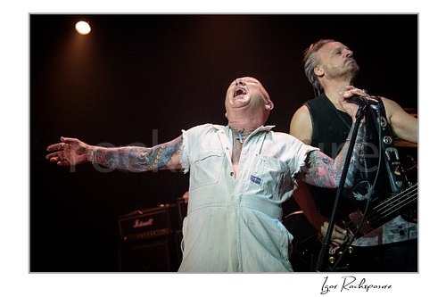 Horizontal color image of Angry Anderson of Rose Tattoo singing onstage with arms outstretched beside a bassist under warm concert light