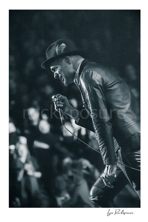 Vertical black and white image of Gord Downie of The Tragically Hip leaning forward with microphone in hand during a live performance, wearing a hat and leather jacket with the crowd softly blurred behind him