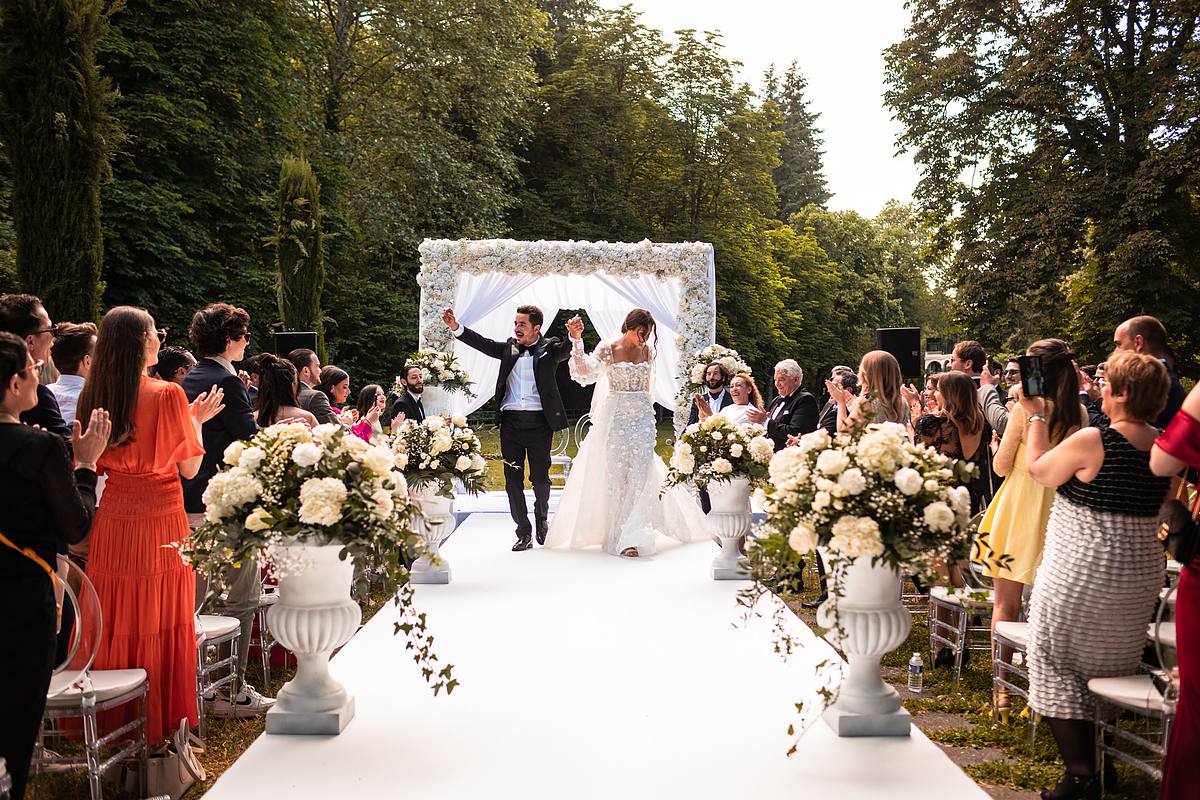 Sebastien CLAVEL Photographe mariage Lyon Le couple de jeunes mari&eacute;s marche main dans la main sur un tapis blanc lors de leur c&eacute;r&eacute;monie de mariage en plein air, entour&eacute; d'invit&eacute;s et de d&eacute;corations florales