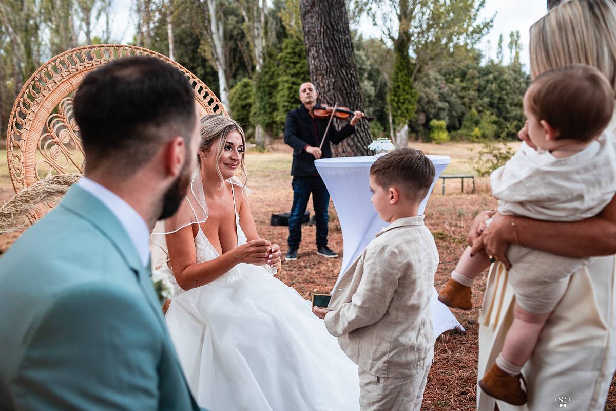 Mari&eacute;e &eacute;changeant un regard complice avec un enfant, violoniste en arri&egrave;re-plan, cr&eacute;ant une ambiance musicale lors d'une c&eacute;r&eacute;monie de mariage &agrave; N&icirc;mes