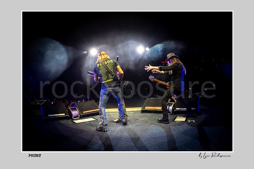 Horizontal color image of Bret Michaels and C.C. DeVille on a Poison stage, photographed with a remote camera mounted on the drum kit facing forward, with bright backlights and stage smoke