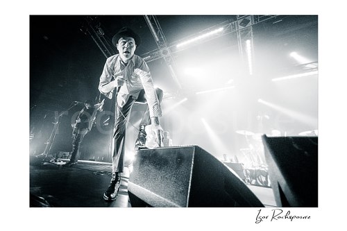 Horizontal black and white image of Gord Downie of The Tragically Hip in a dramatic low-angle wide-lens live performance shot, leaning toward the front of the stage with microphone and white cloth in hand under intense concert light