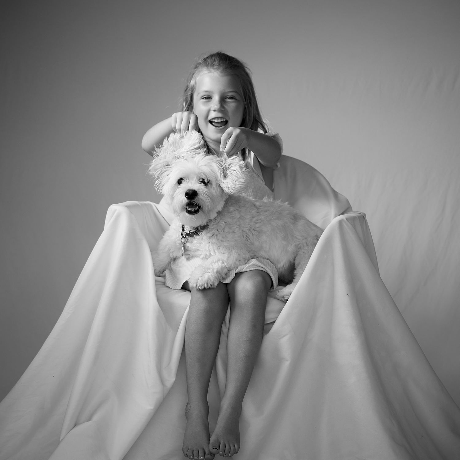 A laughing child playfully holding a small dog&rsquo;s ears in a black-and-white studio portrait