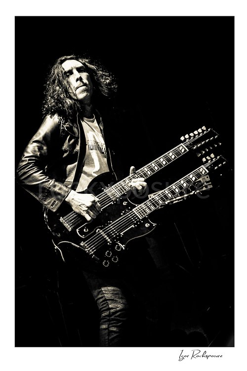 Vertical black and white image of Brent Woods playing double-neck guitar on stage in a dramatic full-body performance shot