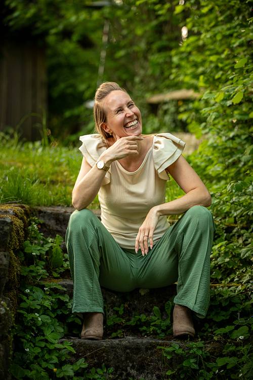 Eine glücklich lachende Frau in einer grünen Hose sitzt auf einer alten, von Pflanzen umgebenen Steintreppe im Freien.
