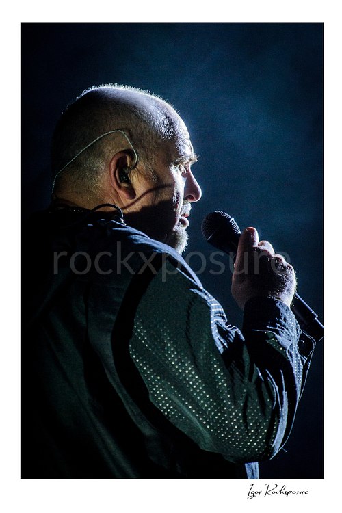 Vertical color image of Peter Gabriel singing into a microphone in side profile under blue concert light