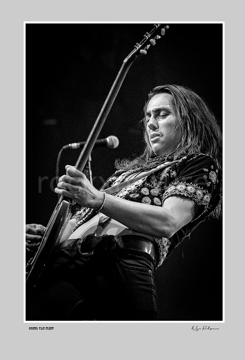 Vertical black and white image of Greta Van Fleet bassist Sam Kiszka performing with his bass guitar on stage