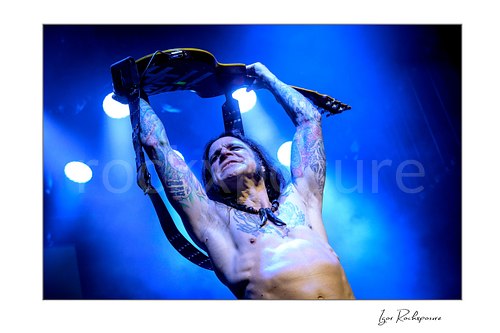 Horizontal color image of Chuck Garric of Beasto Blanco performing live shirtless under vivid blue stage light, raising his guitar overhead with tattooed arms fully extended