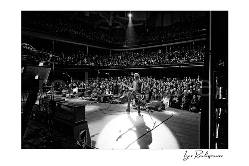 Horizontal black and white image of Nuno Bettencourt standing alone on stage in a spotlight before a packed theater audience