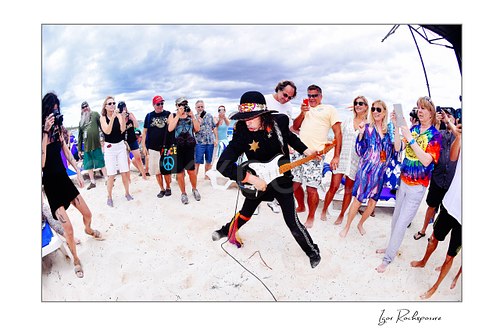 Horizontal color image of Randy Hansen playing guitar on a beach surrounded by cheering fans during an outdoor performance