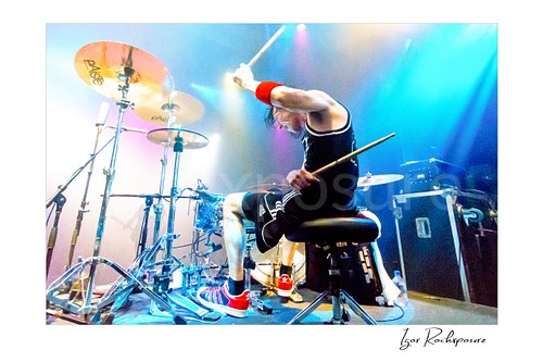 Horizontal color image of Zoltan Chaney playing drums under bright blue and purple stage lighting