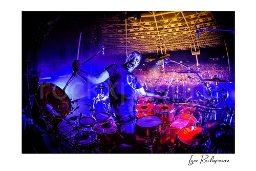 Horizontal color image of Korn drummer Ray Luzier posing at the drums with drumsticks spread wide, a scary facial expression, and the crowd behind him under vivid stage lighting