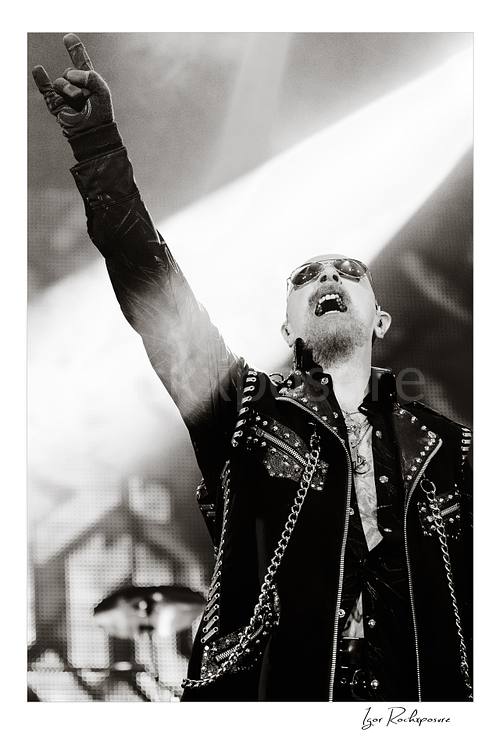 Vertical black and white image of Rob Halford of Judas Priest performing live on stage, raising one arm while holding a microphone under dramatic concert lighting