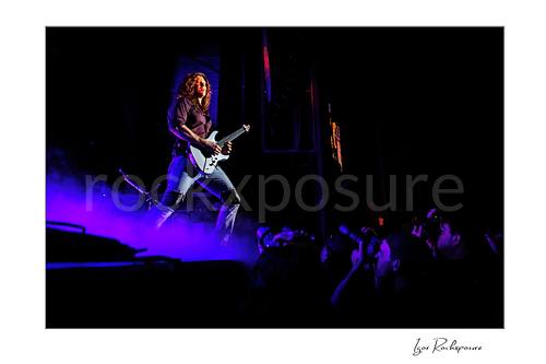Horizontal color image of Kiko Loureiro of Megadeth playing guitar on stage above purple smoke with fans visible below