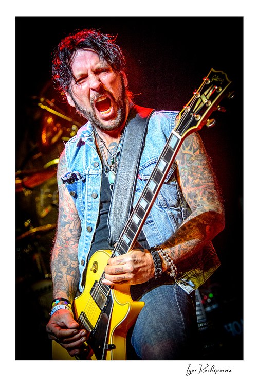 Vertical color image of Tracii Guns performing live with electric guitar in an intense close-up stage portrait