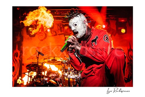 Horizontal color image of Corey Taylor of Slipknot in a red jumpsuit and mask singing into a handheld microphone with fire and the drum kit behind him