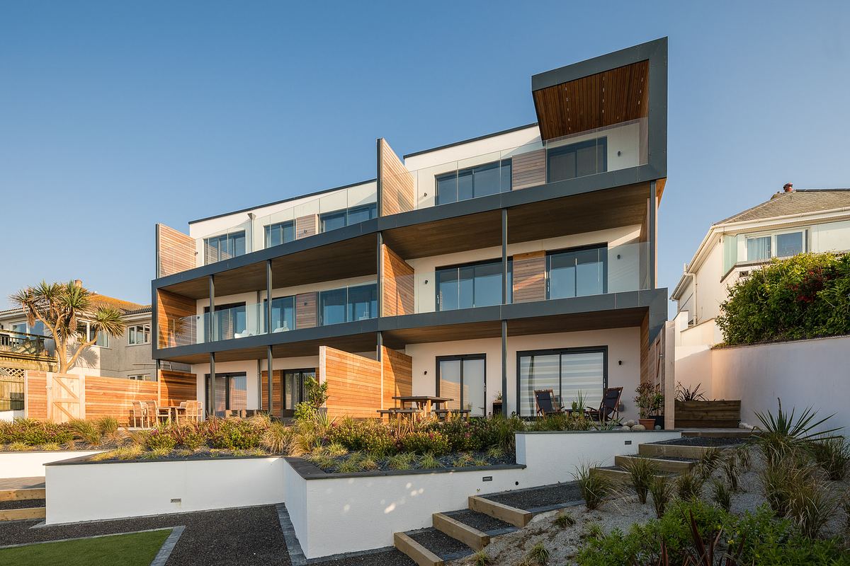 Modern three-story building with balconies and large windows in Cornwall, surrounded by landscaped gardens and situated under a clear sky.