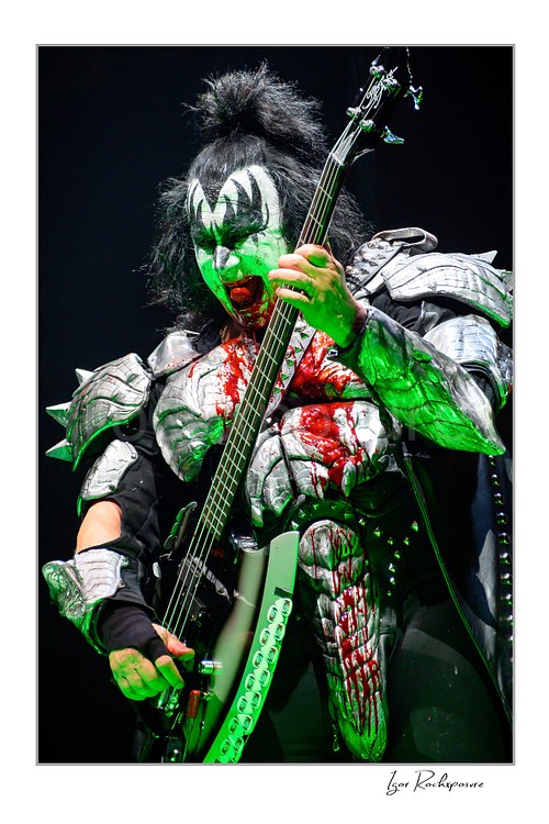 Gene Simmons of KISS in demon makeup covered in stage blood holding his bass guitar under dramatic green concert lighting