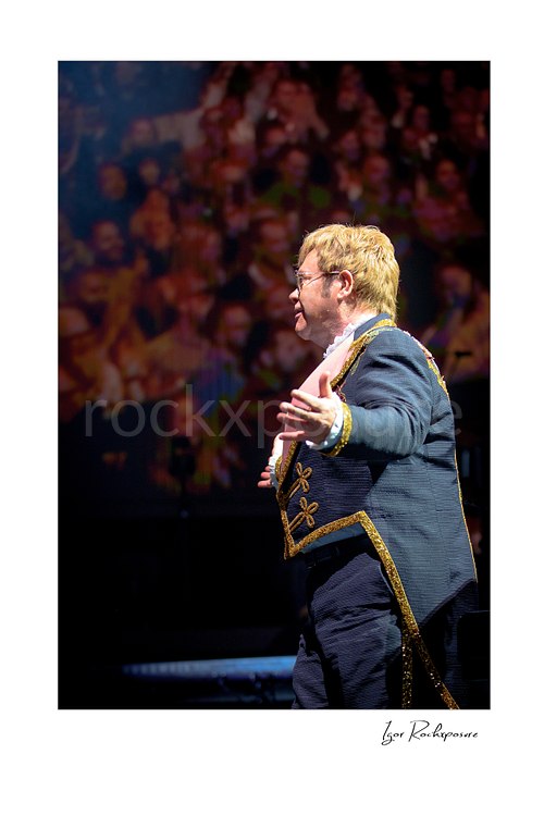 Vertical color image of Elton John on stage in profile with one arm extended before a blurred concert crowd