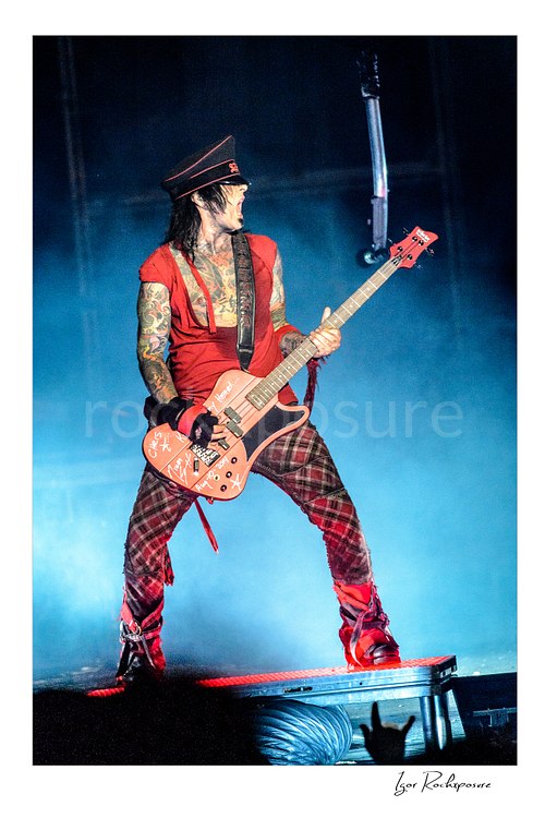 Vertical color image of Nikki Sixx performing with bass guitar during a Mötley Crüe concert in red stage light and blue smoke
