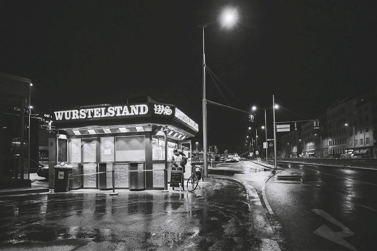 Schwarz-Weiß-Fotografie eines Wiener Würstelstands bei Nacht mit nassem Asphalt, Spiegelungen und urbaner Straßenszene