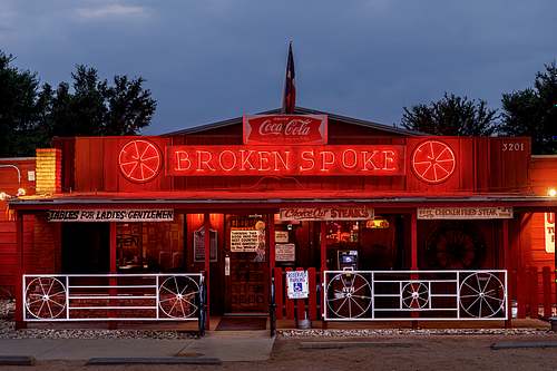 A horizontal fine art color photograph of the iconic Broken Spoke dance hall facade in Austin, Texas, featuring its glowing red neon sign against a twilight sky.