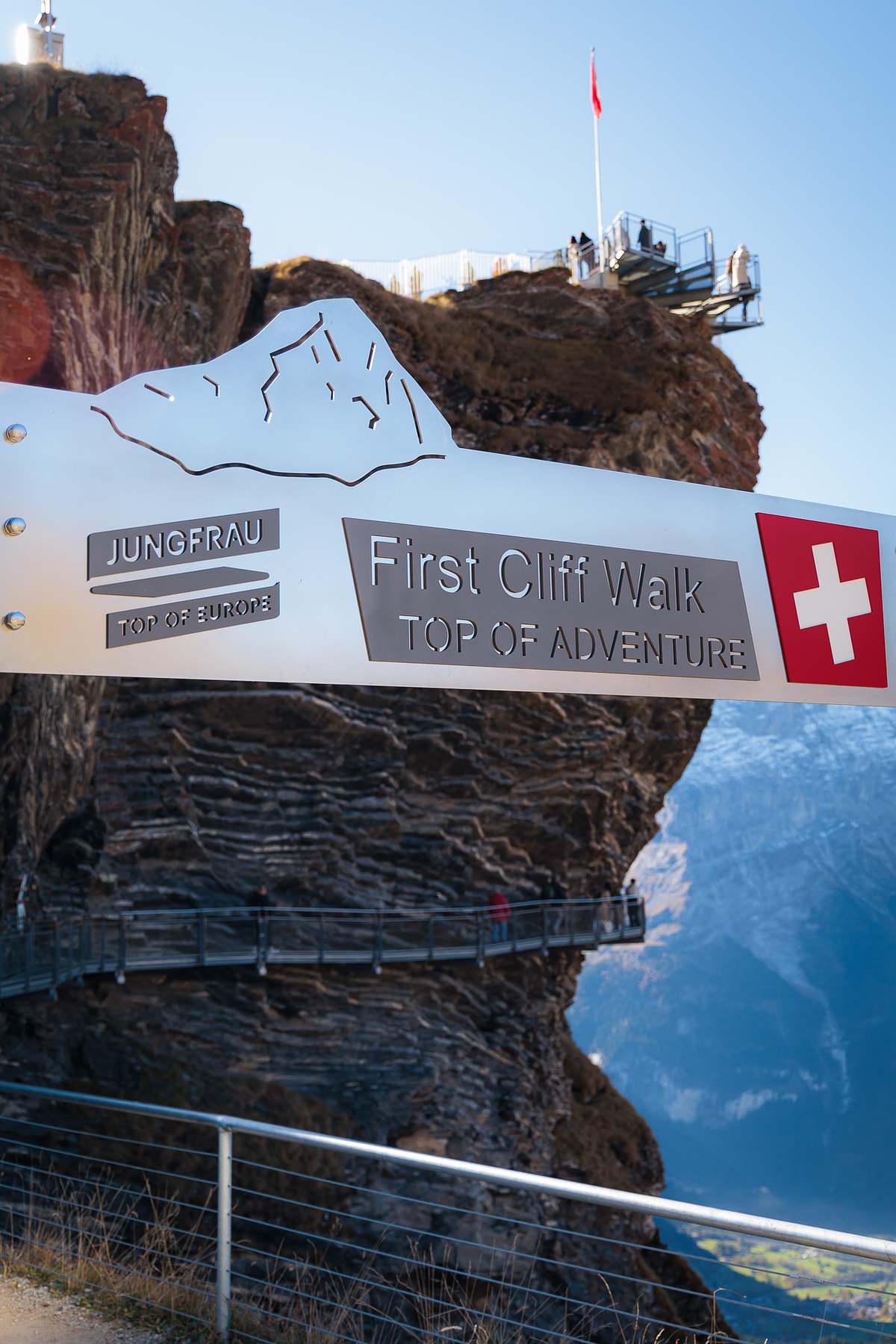 Geb&uuml;rstetes Metallschild mit Schriftzug First Cliff Walk Top of Adventure und Schweizer Kreuz, dahinter Wanderer auf dem in den Fels gebauten Steg und eine Aussichtsplattform mit Schweizer Flagge