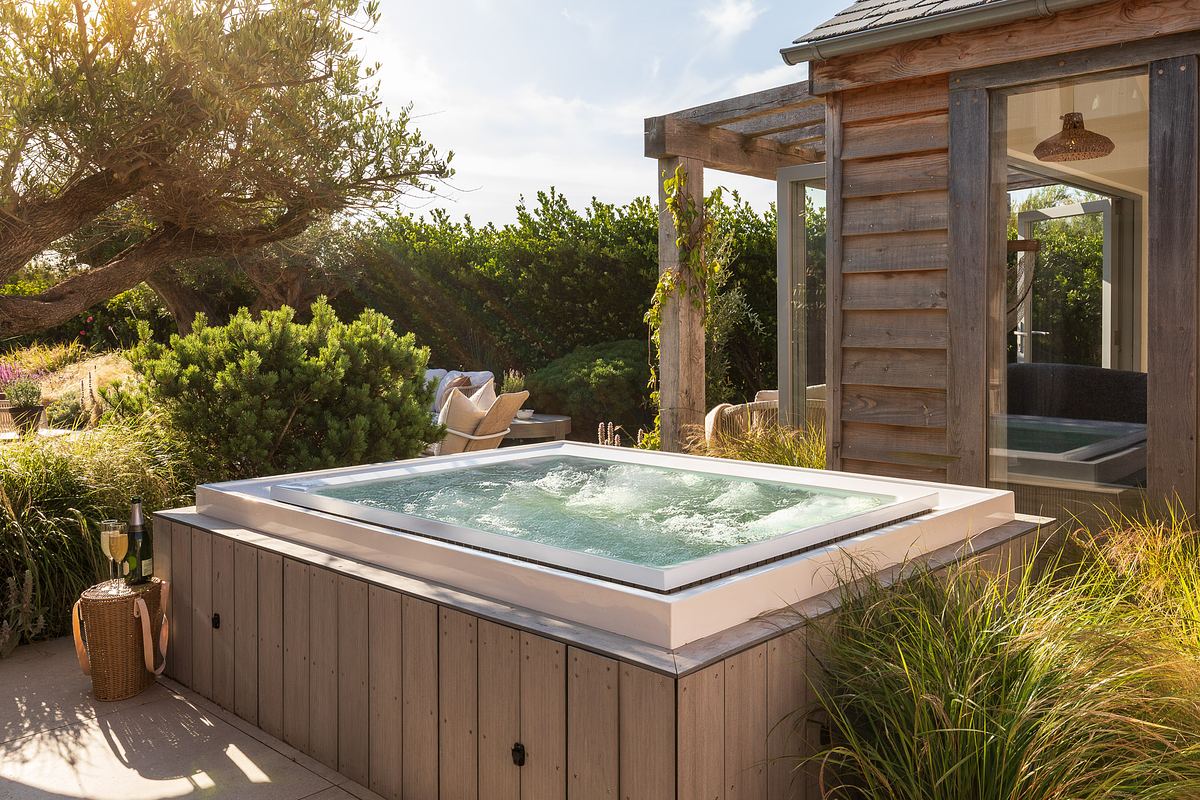 A modern square hot tub with bubbling water is set on a wooden deck surrounded by green plants, next to a rustic summer house with large windows.