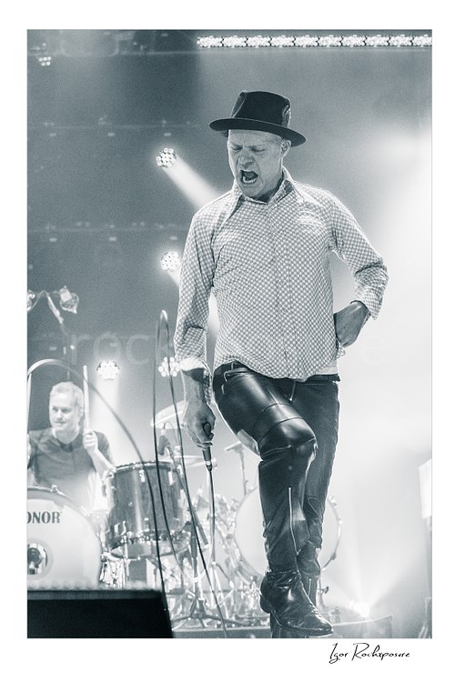 Vertical black and white image of Gord Downie of The Tragically Hip in a dramatic live performance pose, stepping across the stage with microphone in hand while Johnny Fay plays drums behind him