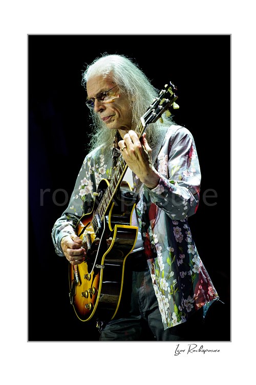 Vertical color image of Steve Howe of Yes playing guitar onstage in glasses and a floral shirt during a live performance