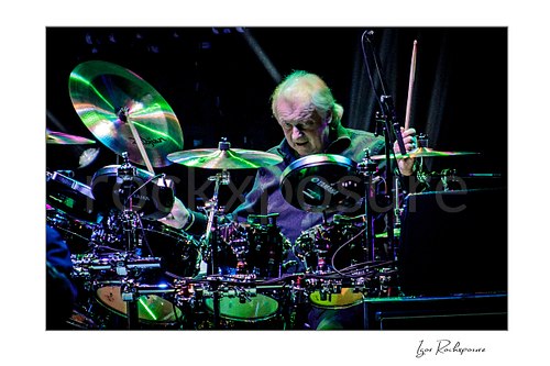 Horizontal color image of Alan White of Yes performing at the drum kit under vivid green concert lighting