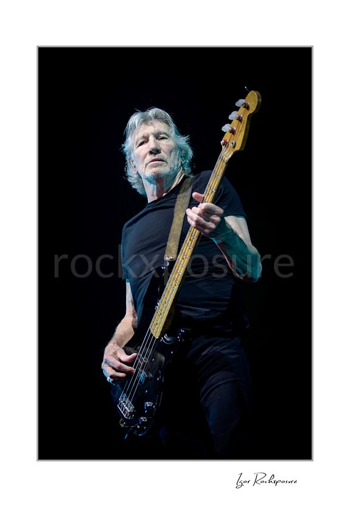 Roger Waters looks outward while playing a black bass with its wooden neck angled upward, illuminated by cool light against a black stage.