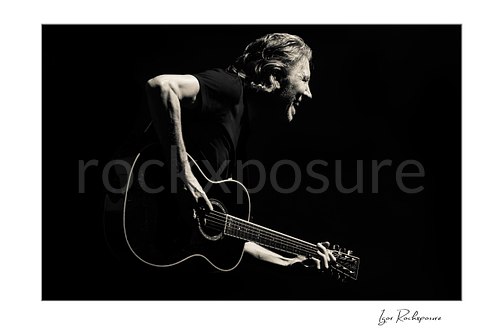 Black and white photograph of Roger Waters smiling and leaning forward while playing a dark acoustic guitar against a nearly black stage background.