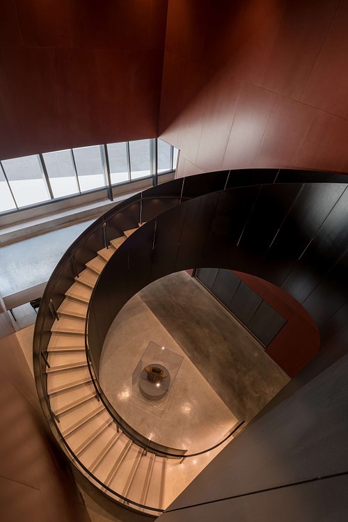 Looking down a dramatic spiral staircase at the Winter Park Events Center in Florida, designed by architect David Adjaye, showcasing modern geometric curves and warm wood tones.