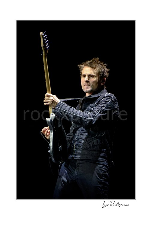 Vertical color image of Matt Bellamy of Muse holding guitar upright in a dramatic stage portrait against a dark background