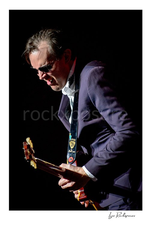 Vertical color image of Joe Bonamassa performing live on stage with a guitar in a dramatic close concert portrait