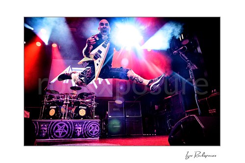 Horizontal color image of Scott Ian of Anthrax performing live on stage, jumping with his guitar under vivid red and blue concert lighting