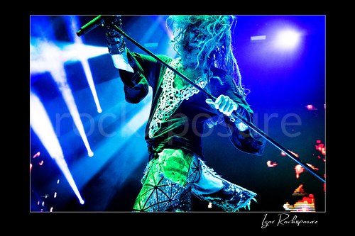 Horizontal color image of Rob Zombie performing live on stage under blue lighting with flying dreadlocks, a tilted microphone stand, and reflective stagewear