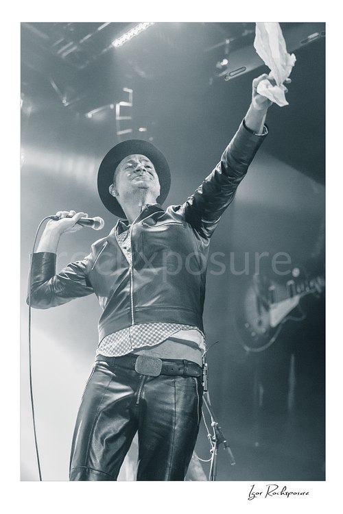 Vertical black and white image of Gord Downie of The Tragically Hip holding a microphone and lifting a white cloth high on stage, wearing a hat and leather jacket in bright concert haze