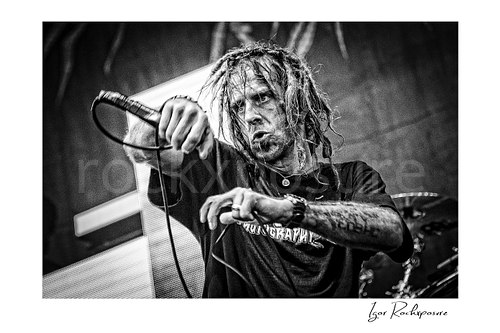 Horizontal black and white image of Randy Blythe of Lamb of God performing on stage with dreadlocks, handheld microphone, and an intense expression