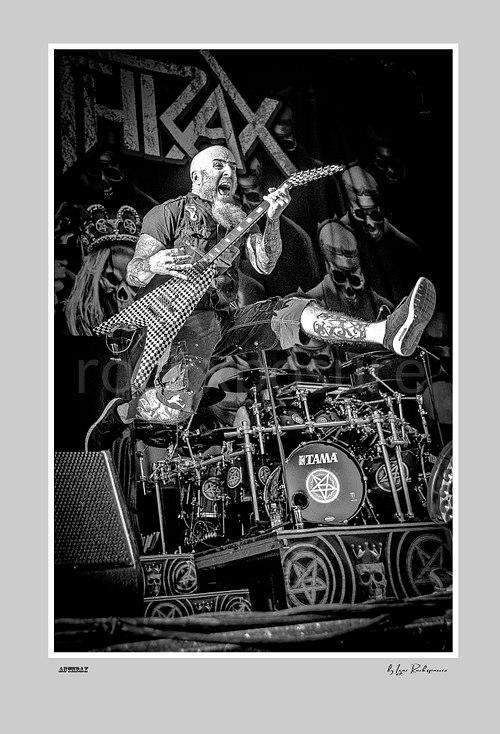 Vertical black and white image of Scott Ian of Anthrax performing live on stage, jumping with his guitar in front of the drum kit and skull-themed stage backdrop