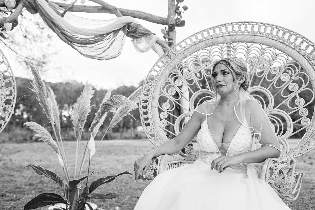La mari&eacute;e tres &eacute;mue et en pleurs s'assoit majestueusement sur un fauteuil peacock en rotin lors de sa c&eacute;r&eacute;monie de mariage &agrave; N&icirc;mes, encadr&eacute;e par des ornements floraux et le paysage naturel, une sc&egrave;ne capturant l'&eacute;l&eacute;gance rustique et la s&eacute;r&eacute;nit&eacute;