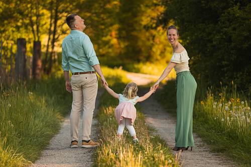 Vater, Tochter und Mutter stehen an den Händen haltend auf einem Feldweg und lachen gemeinsam in der Natur.