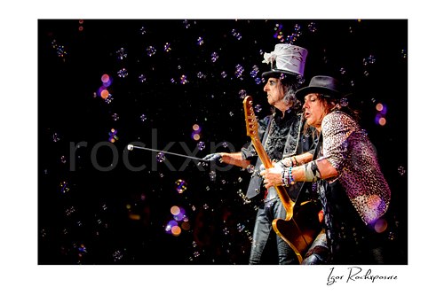 Horizontal color image of Alice Cooper and Ryan Roxie performing live on stage with a silver top hat, guitar, cane, and bubbles floating through the air