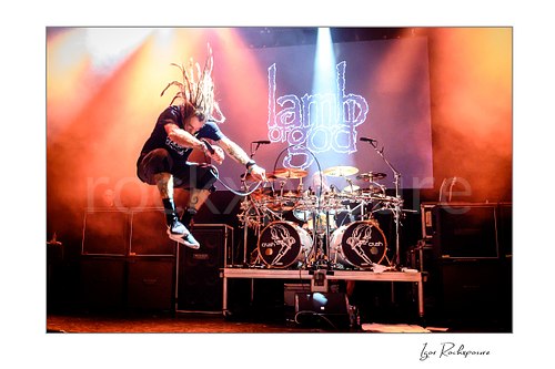 Horizontal color image of Randy Blythe of Lamb of God leaping on stage with raised dreadlocks, handheld microphone, and the drum kit behind him