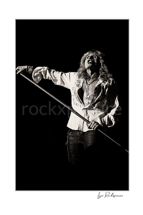 Vertical black and white image of David Coverdale of Whitesnake performing live on stage, holding a microphone stand against a dark background