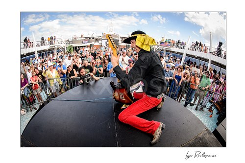 Horizontal color image of Randy Hansen kneeling on an outdoor ship deck stage with guitar and a large crowd behind him