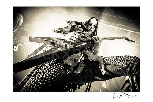 Horizontal black and white image of Nergal of Behemoth playing guitar in an intense low-angle close-up under bright stage backlight