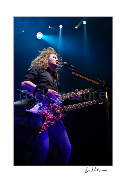Vertical color image of Dave Mustaine of Megadeth singing and playing a double-neck guitar on stage under blue spotlights